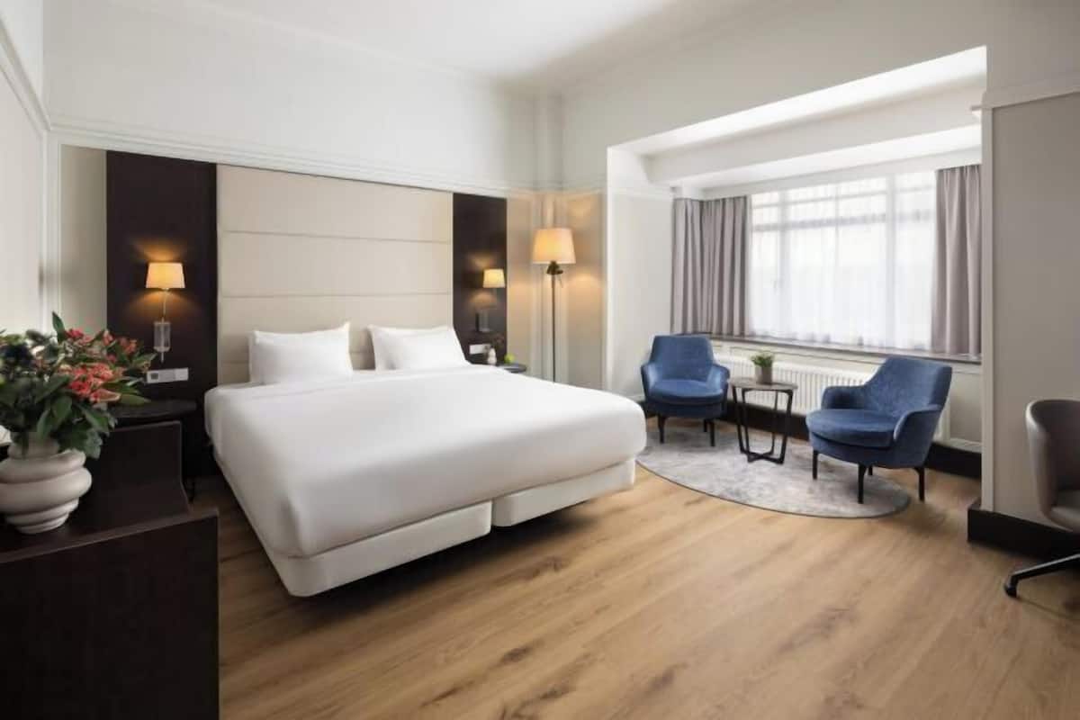 A hotel room with a large bed, two armchairs, a small table, and a window with curtains.