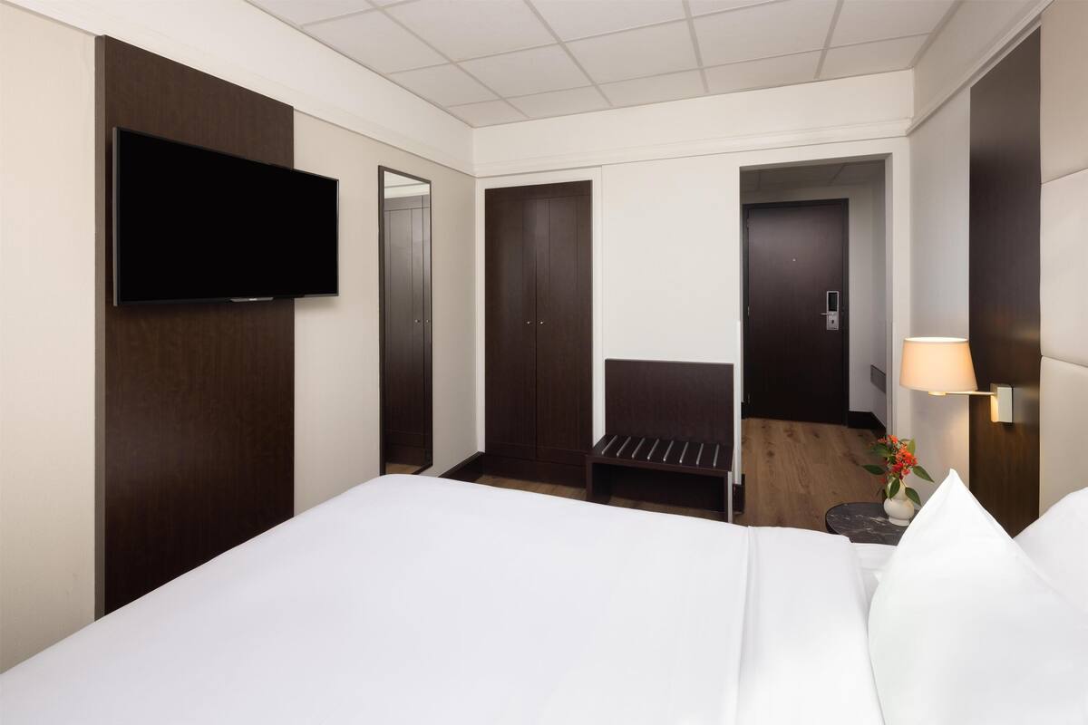 A hotel room with a bed, a flat-screen TV mounted on the wall, a wooden bench, a small table with a lamp, and a door leading to another room.
