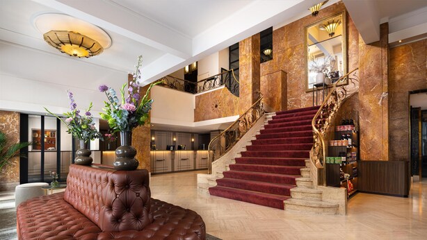 A grand lobby with a red carpeted staircase, a tufted leather sofa, and a large vase of flowers. - NH Atlanta Rotterdam (Rotterdam)