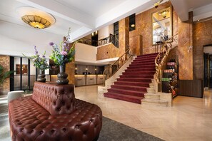 A grand lobby with a red carpeted staircase, a tufted leather sofa, and a large vase of flowers.
