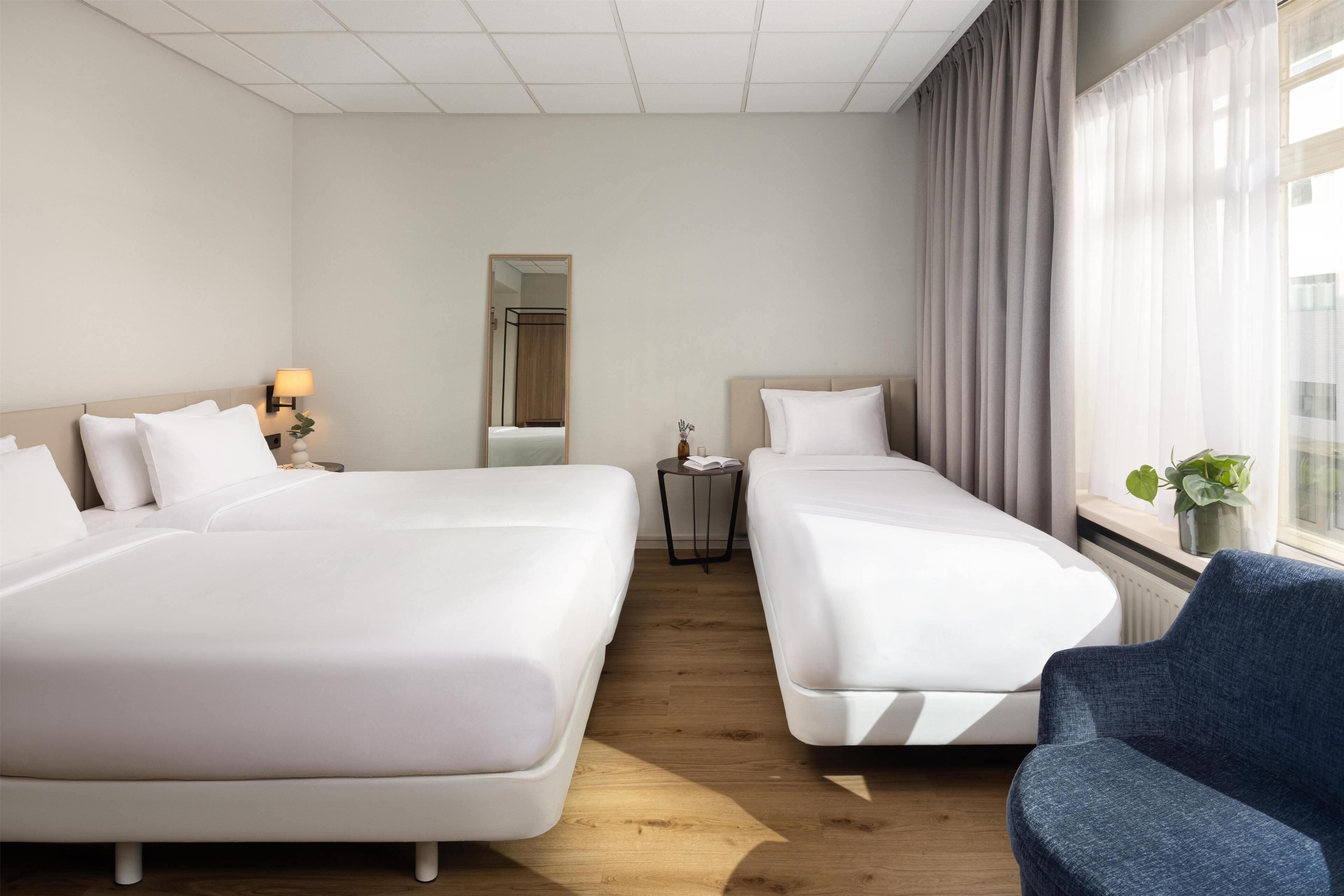 Habitación de hotel con dos camas, una mesita con lámpara, un espejo y una ventana con cortinas.