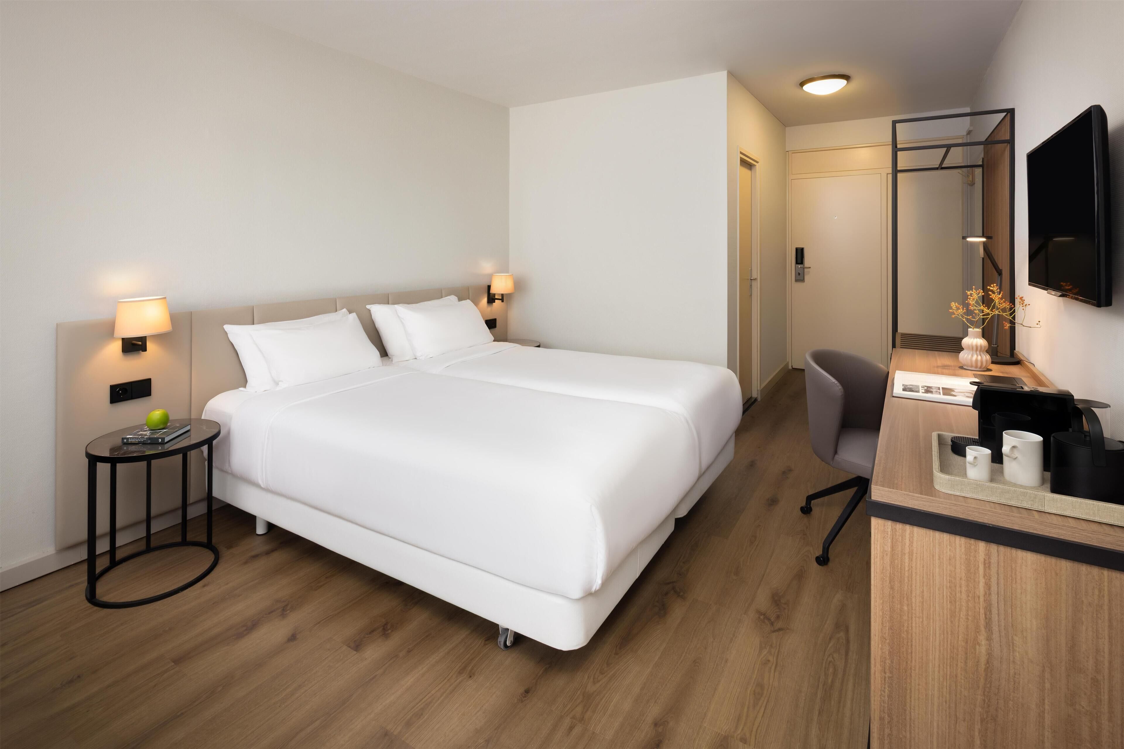 A hotel room with a large bed, bedside lamps, a desk with a chair, a mirror, and a flat-screen TV.