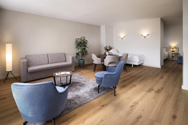 A modern living room with a grey sofa, blue armchairs, a coffee table, and a dining table with chairs. There is a potted plant and a floor lamp.