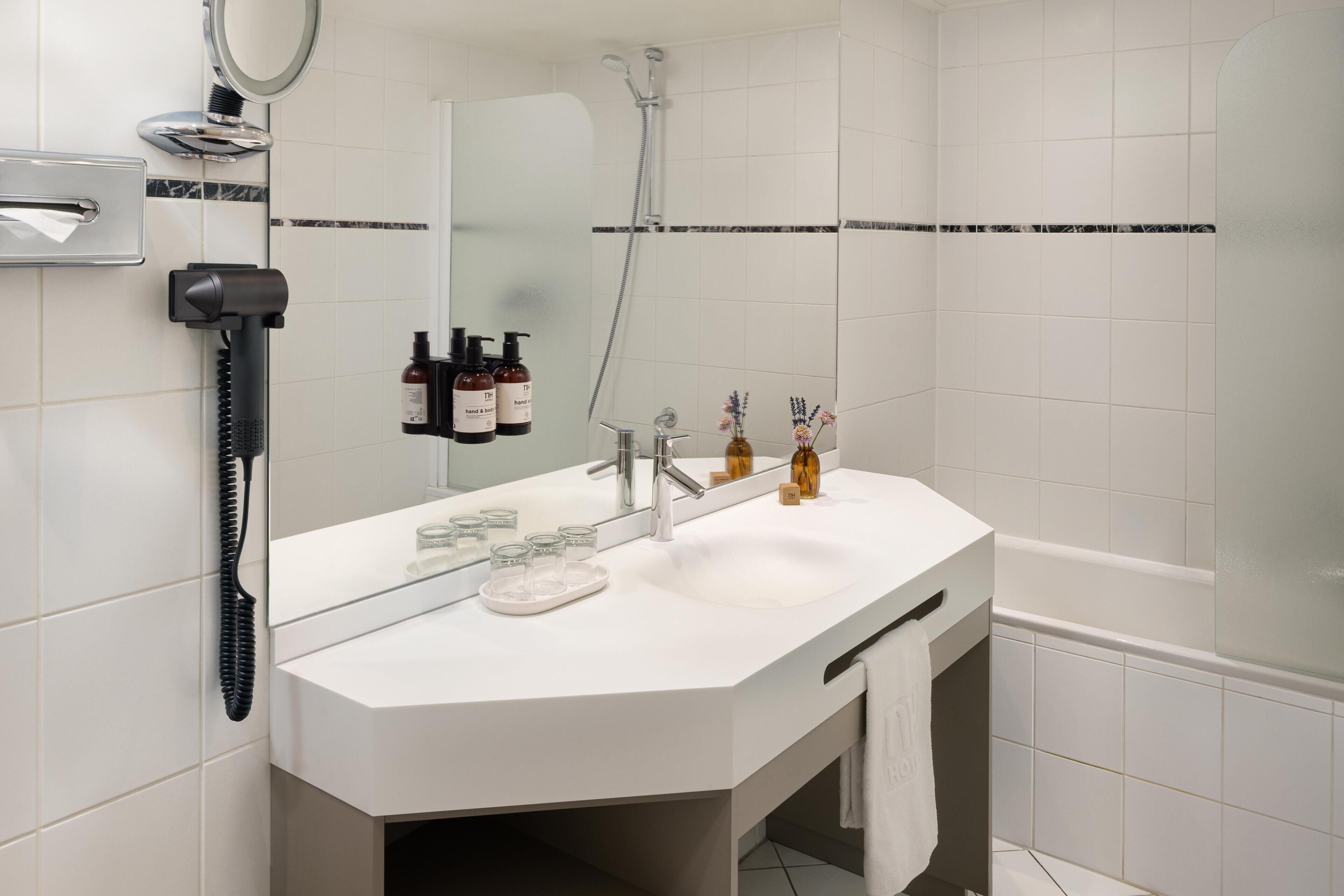 Un baño con lavamanos blanco, espejo y ducha. Hay artículos de aseo en la pared y un perchero.