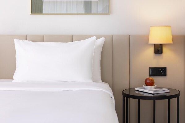 A neatly made bed with white bedding and pillows, a round black side table with a lamp and a book, and a framed picture on the wall.