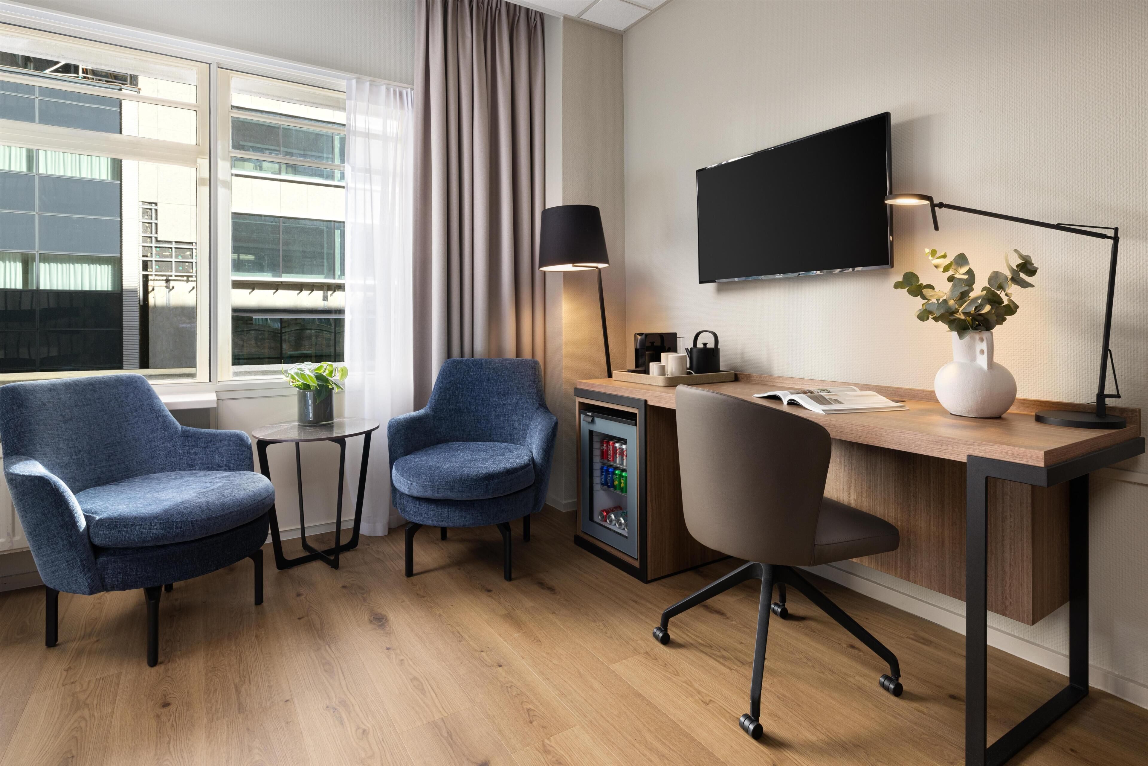 Habitación de hotel moderna con un escritorio de madera, dos sillones azules, una mesita con una planta, un televisor montado en la pared y un refrigerador minibar.