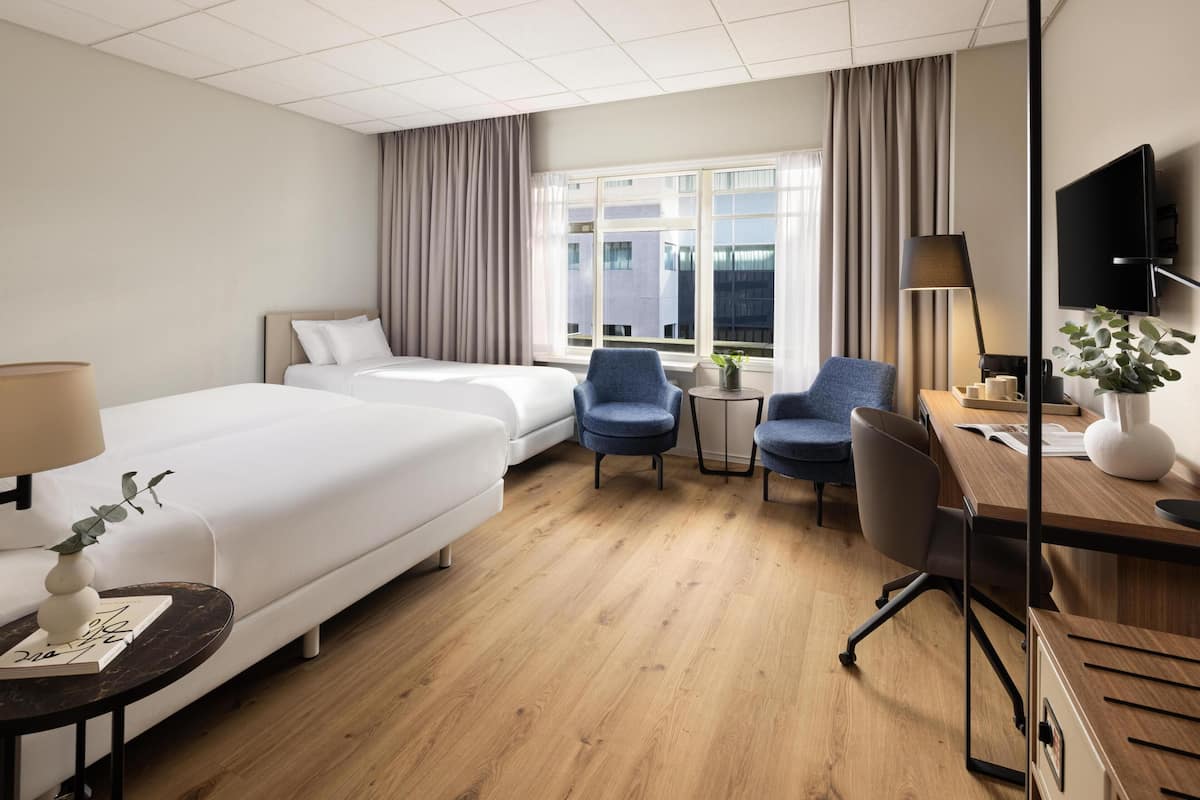 A hotel room with two beds, a desk, a chair, a TV, and a window with curtains.