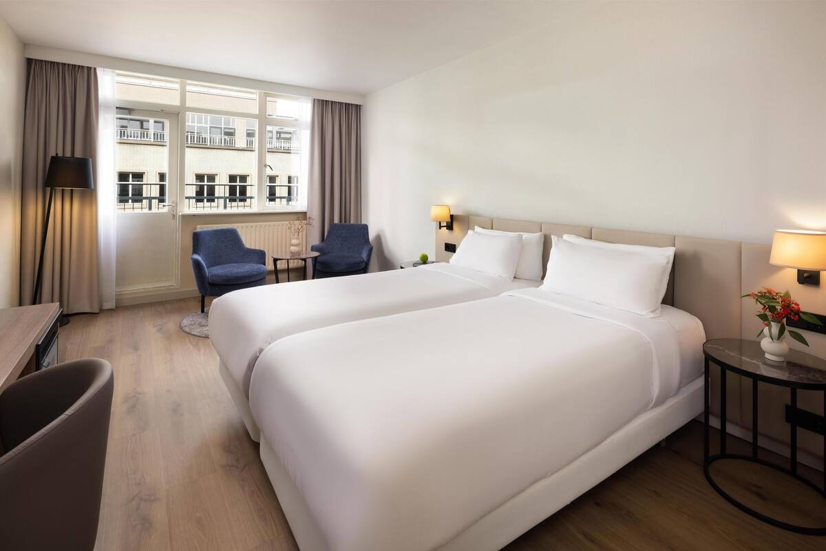 A hotel room with two beds, a desk, a chair, a window with curtains, and a view of a building.