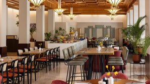 A spacious dining area with high ceilings, multiple tables, and chairs, featuring a central island with a fruit display and a potted plant. - NH Atlanta Rotterdam (Rotterdam)