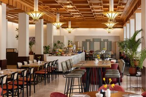 A spacious dining area with high ceilings, multiple tables, and chairs, featuring a central island with a fruit display and a potted plant. - NH Atlanta Rotterdam (Rotterdam)