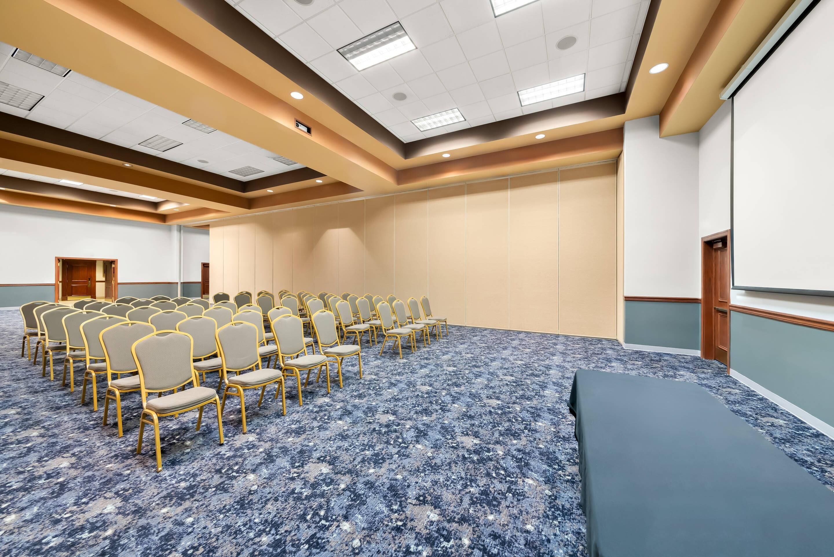 A conference room with rows of chairs, a carpeted floor, a projector screen, and a stage area.