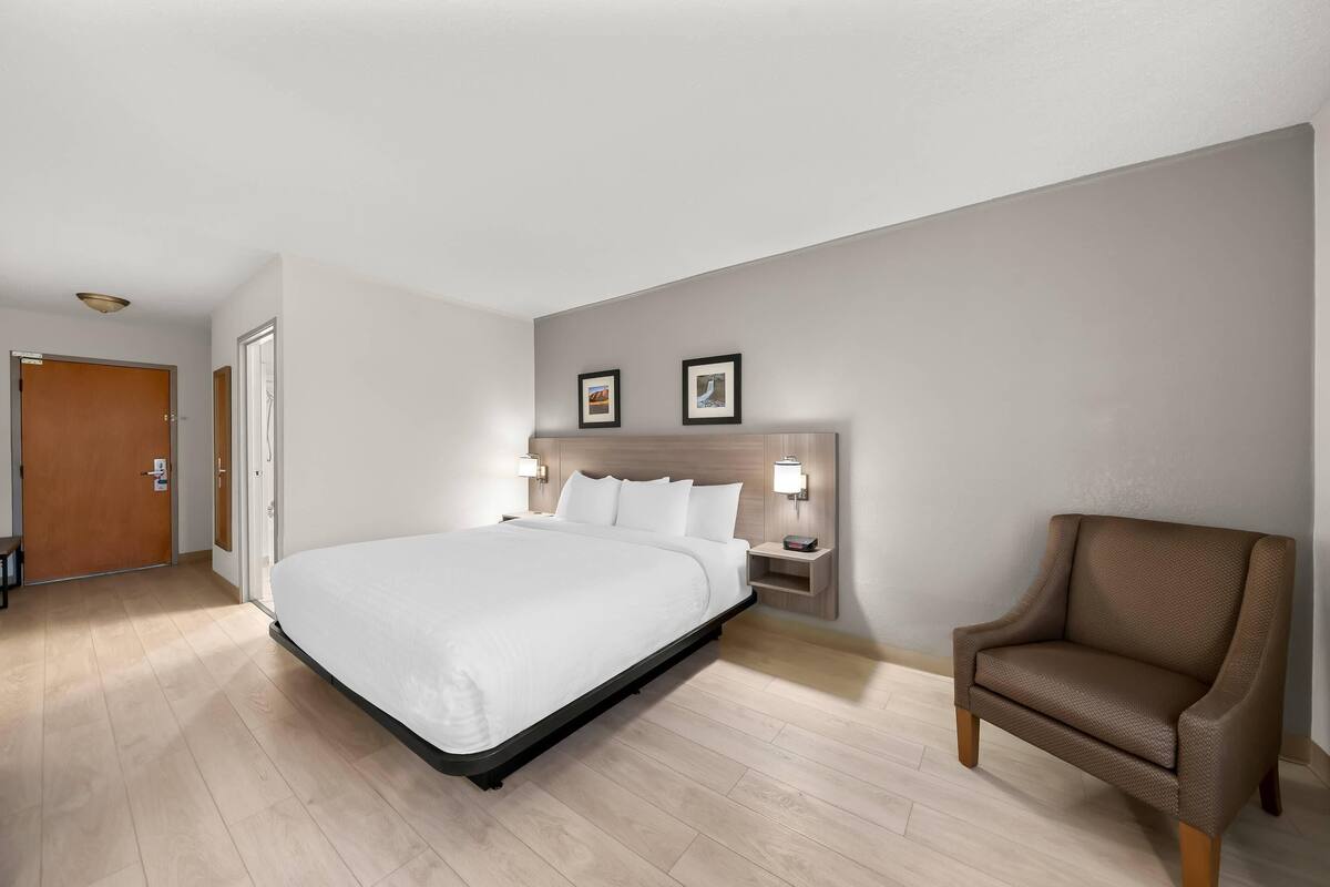 A hotel room with a bed, a chair, a nightstand with a lamp, and a door.