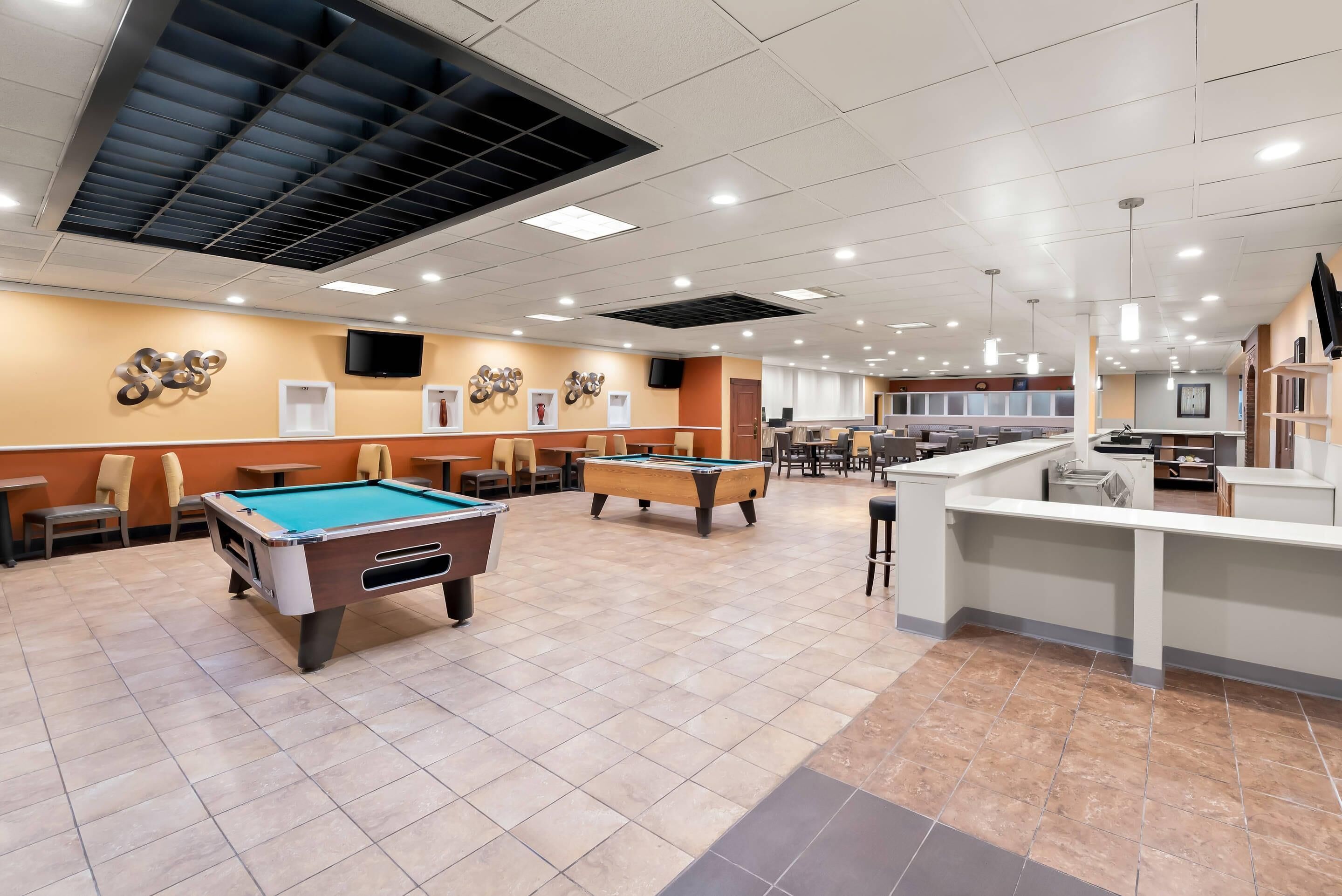 A pool table, a billiard table, and a bar area in a well-lit indoor space with ceiling lights and a flat-screen TV mounted on the wall.