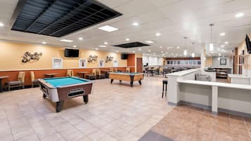 A pool table, a billiard table, and a bar area in a well-lit indoor space with ceiling lights and a flat-screen TV mounted on the wall.