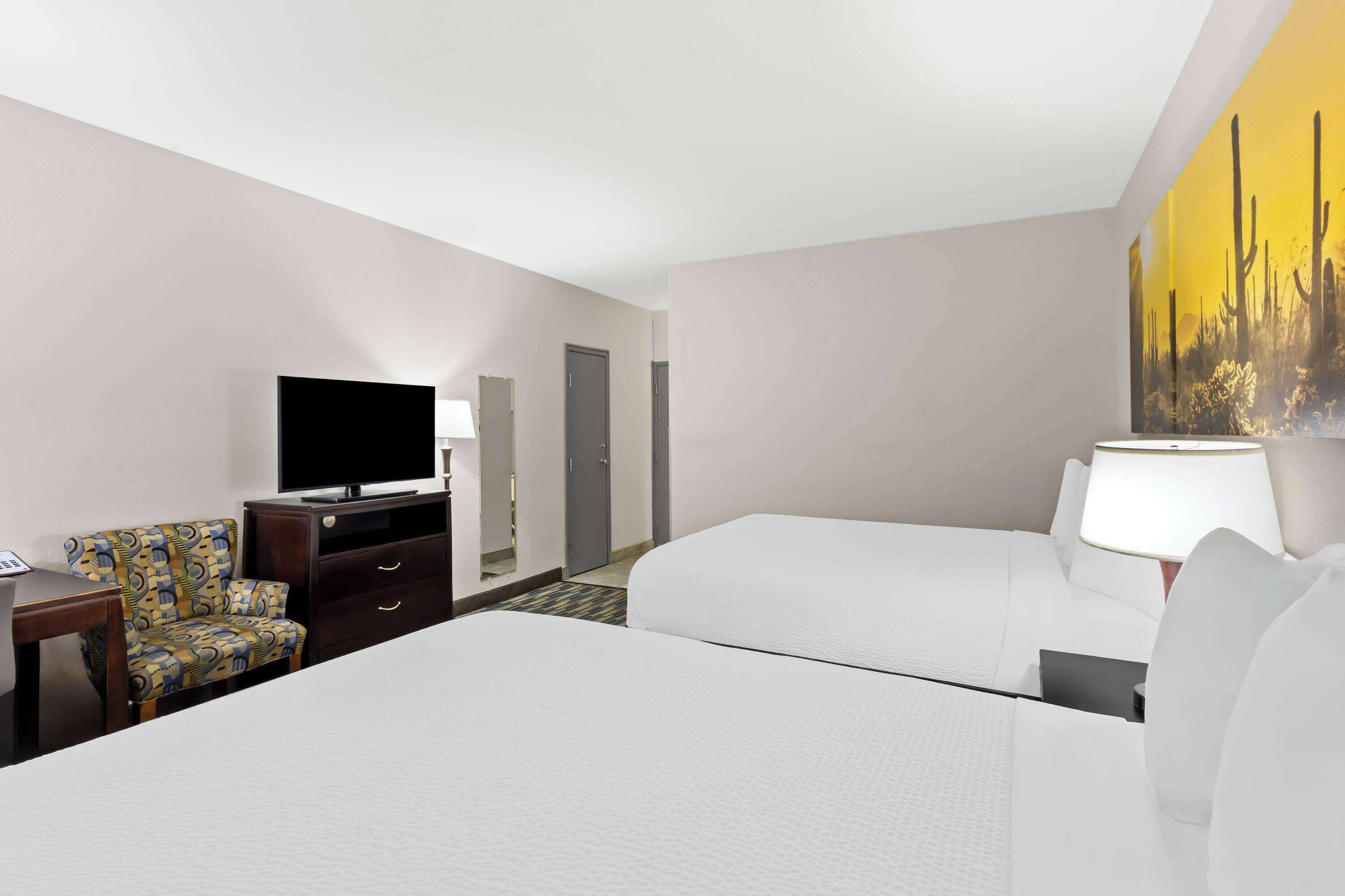 A hotel room with a bed, a TV, a desk, a chair, and a painting on the wall.