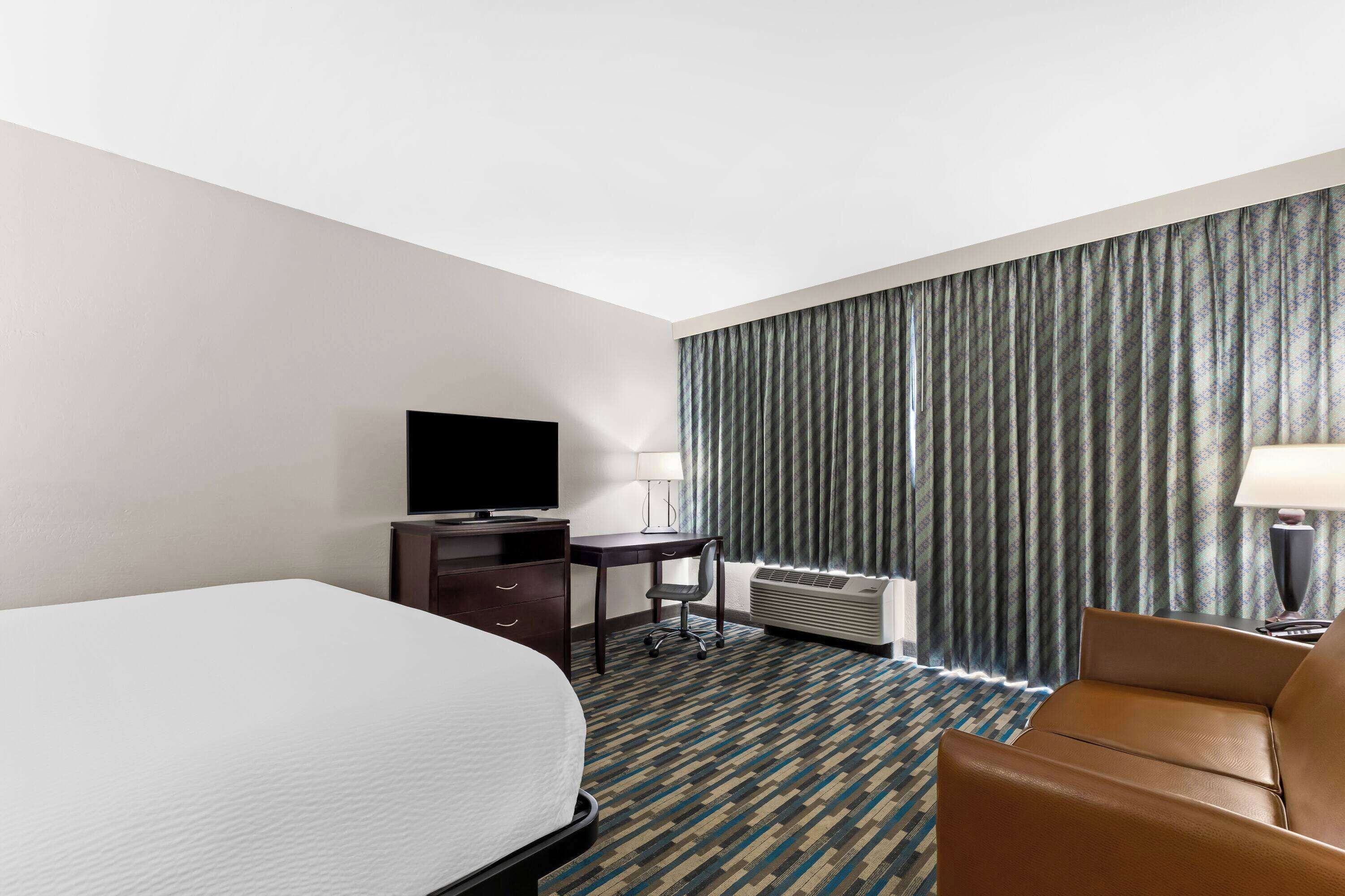 Habitación de hotel con cama, escritorio, silla, televisor y ventana con cortinas.