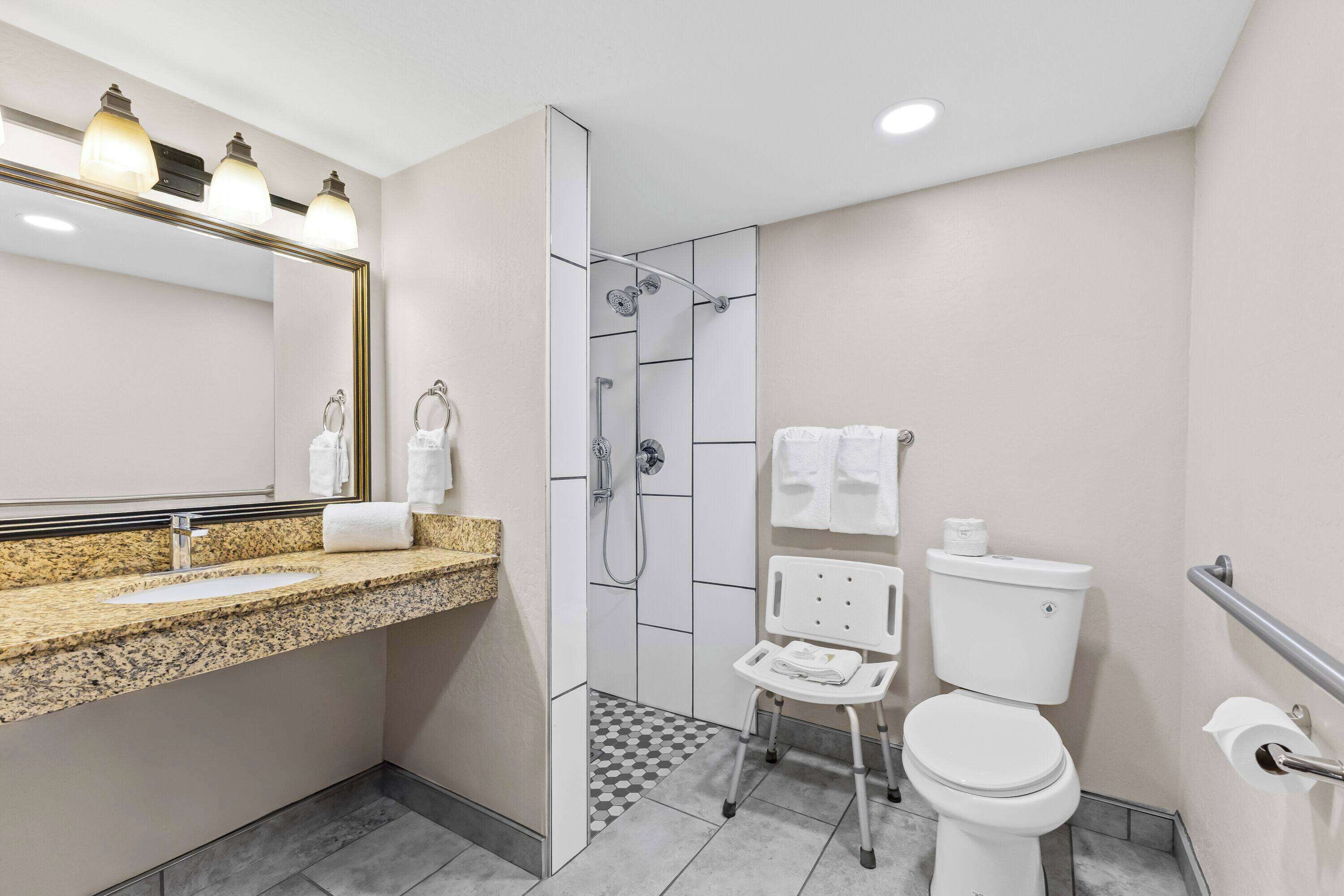 Une salle de bain moderne avec un comptoir en granit, une cabine de douche en verre, des toilettes blanches et une barre d’appui.
