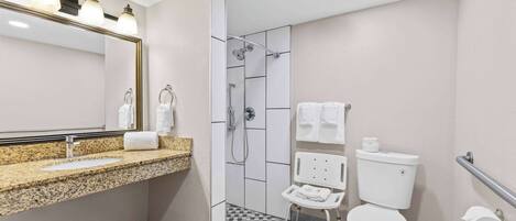 Une salle de bain moderne avec un comptoir en granit, une cabine de douche en verre, des toilettes blanches et une barre d’appui.
