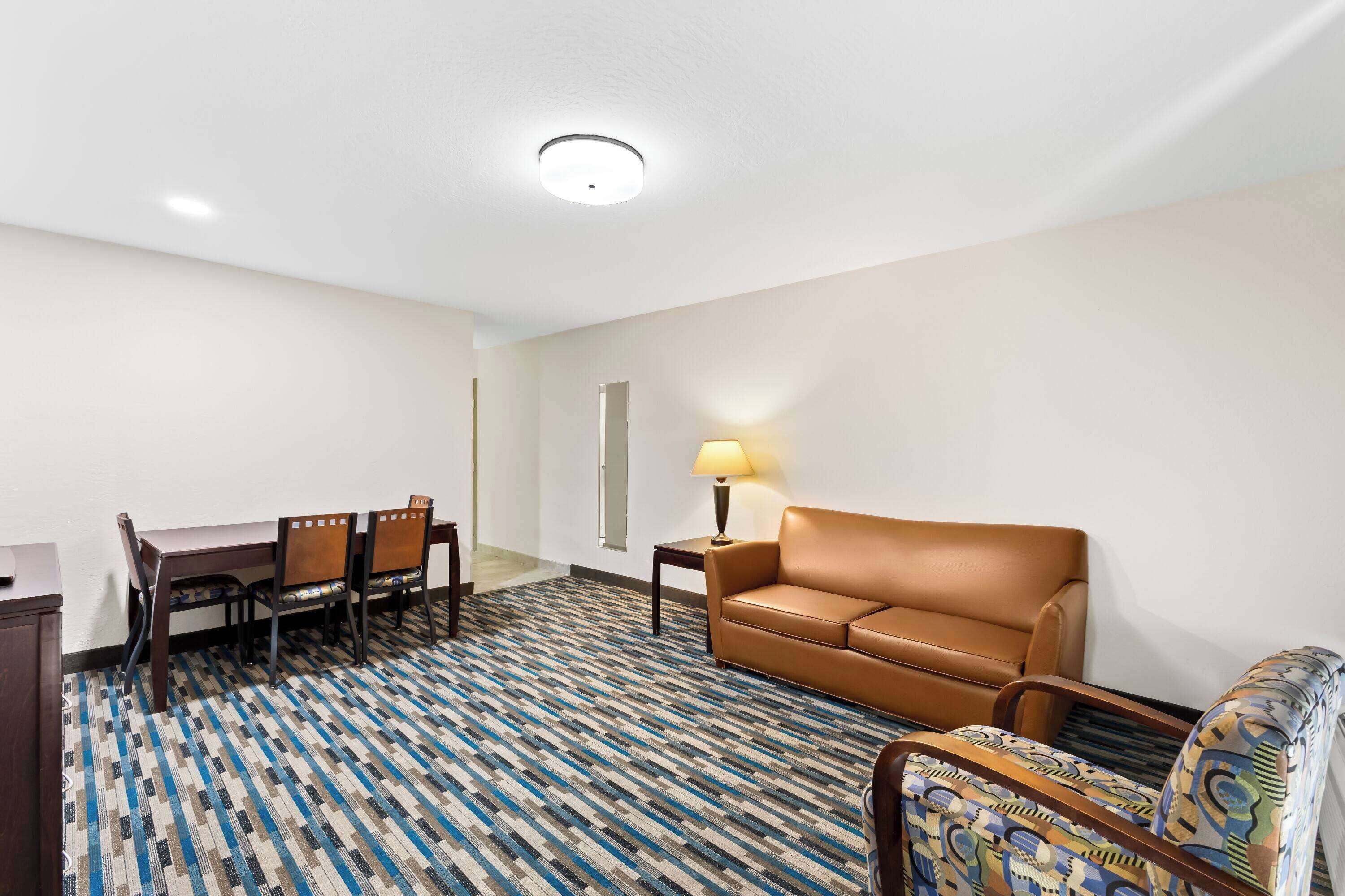 Habitación de hotel con alfombra estampada, sofá de cuero marrón, mesa de comedor con sillas y una lámpara sobre una mesita.