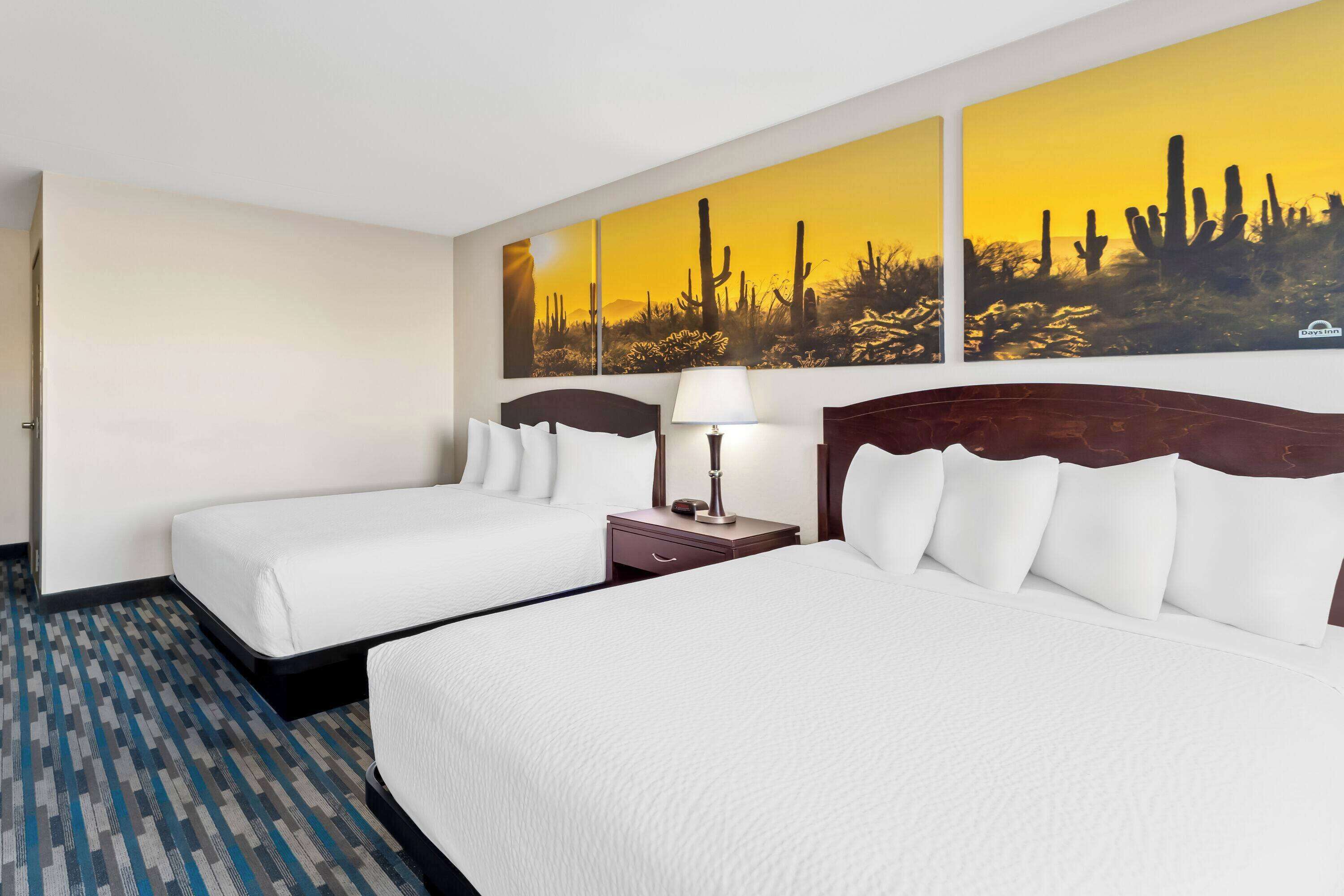 Habitación de hotel con dos camas, una mesita de noche con lámpara y un cuadro con temática desértica.