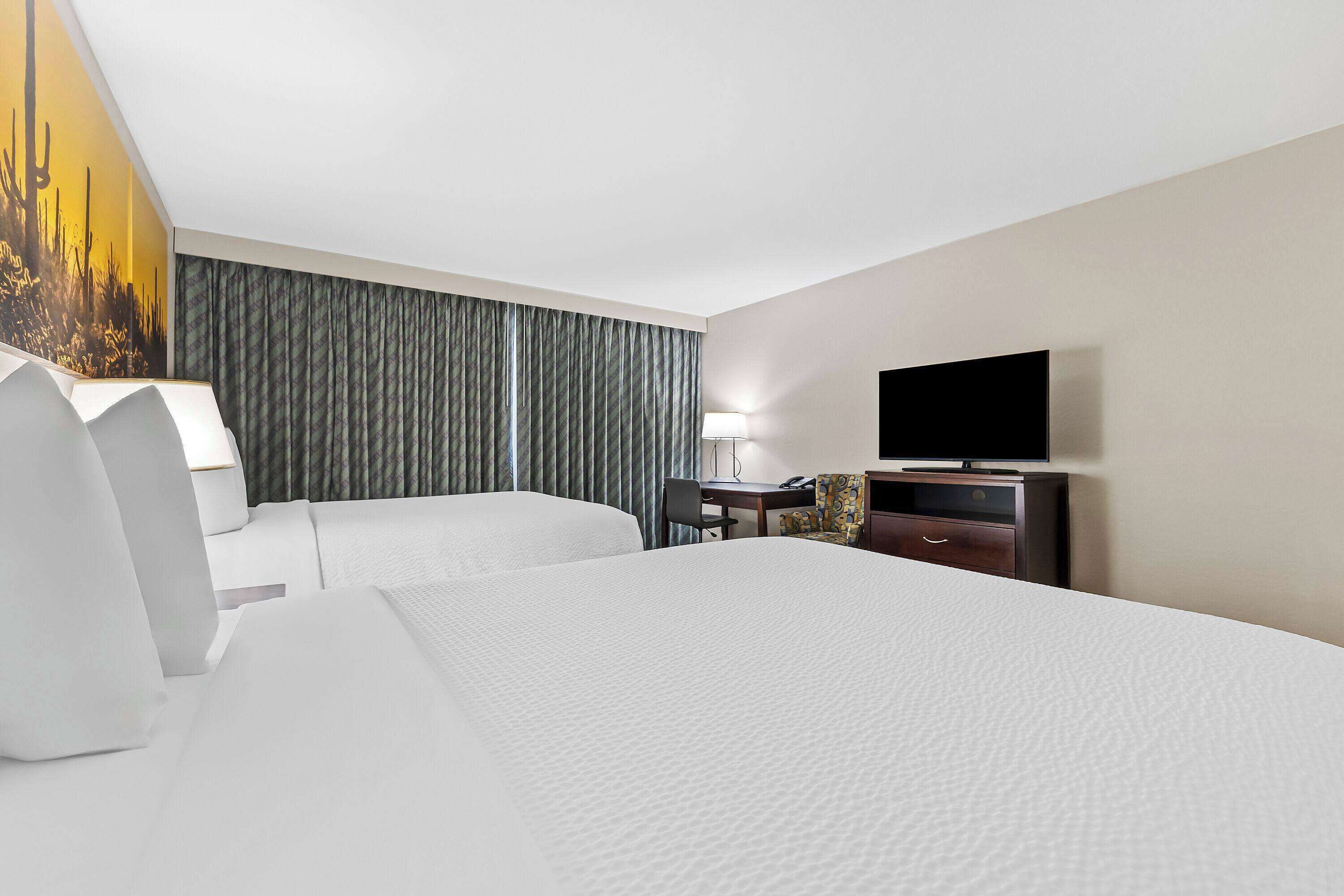 A hotel room with two beds, a desk with a chair, a television, and a large window with curtains.