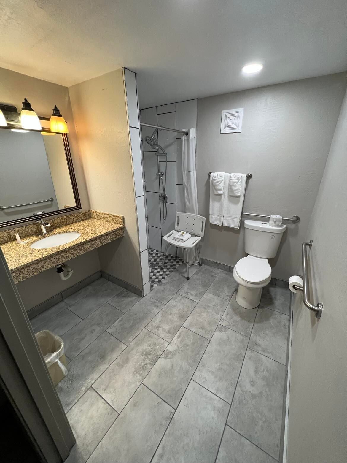 Un baño moderno con lavamanos de granito, ducha, inodoro y una pequeña mesa con silla.