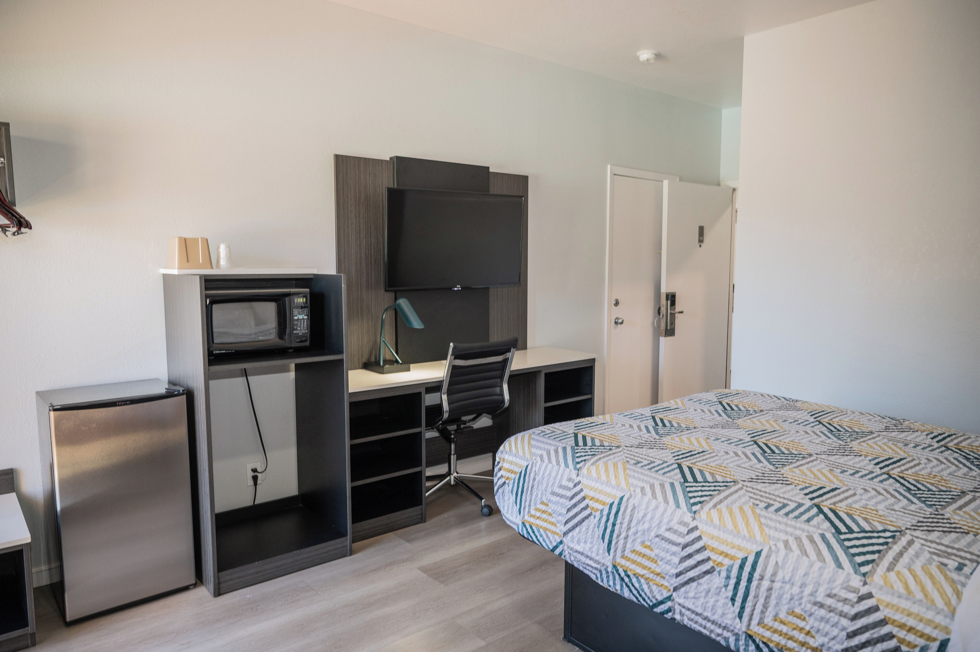 A compact hotel room with a bed, a desk with a computer and a microwave, and a small refrigerator.