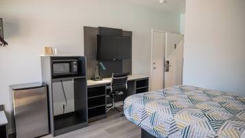 A compact hotel room with a bed, a desk with a computer and a microwave, and a small refrigerator.