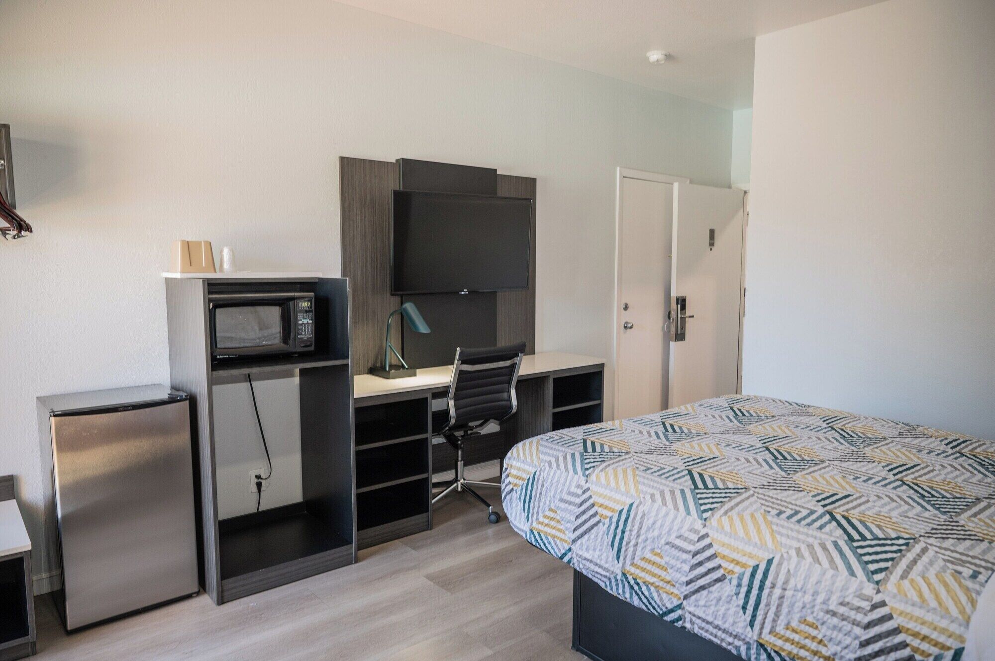 Habitación de hotel pequeña con kitchenette, una cama con cubrecama estampado, un escritorio con silla y un televisor montado en la pared.