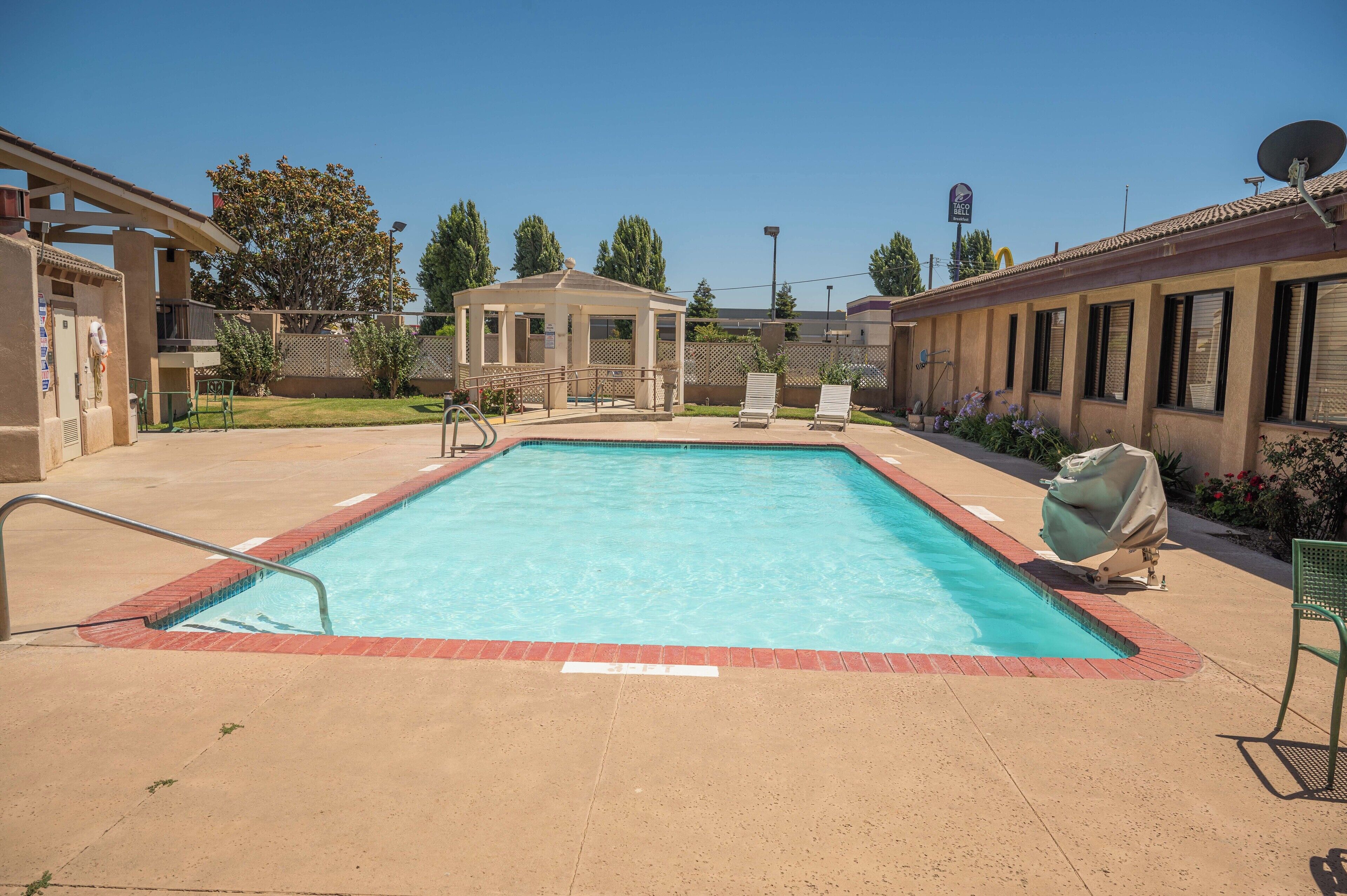 A rectangular swimming pool with a red border, surrounded by a concrete deck. There are lounge chairs and a covered grill area.