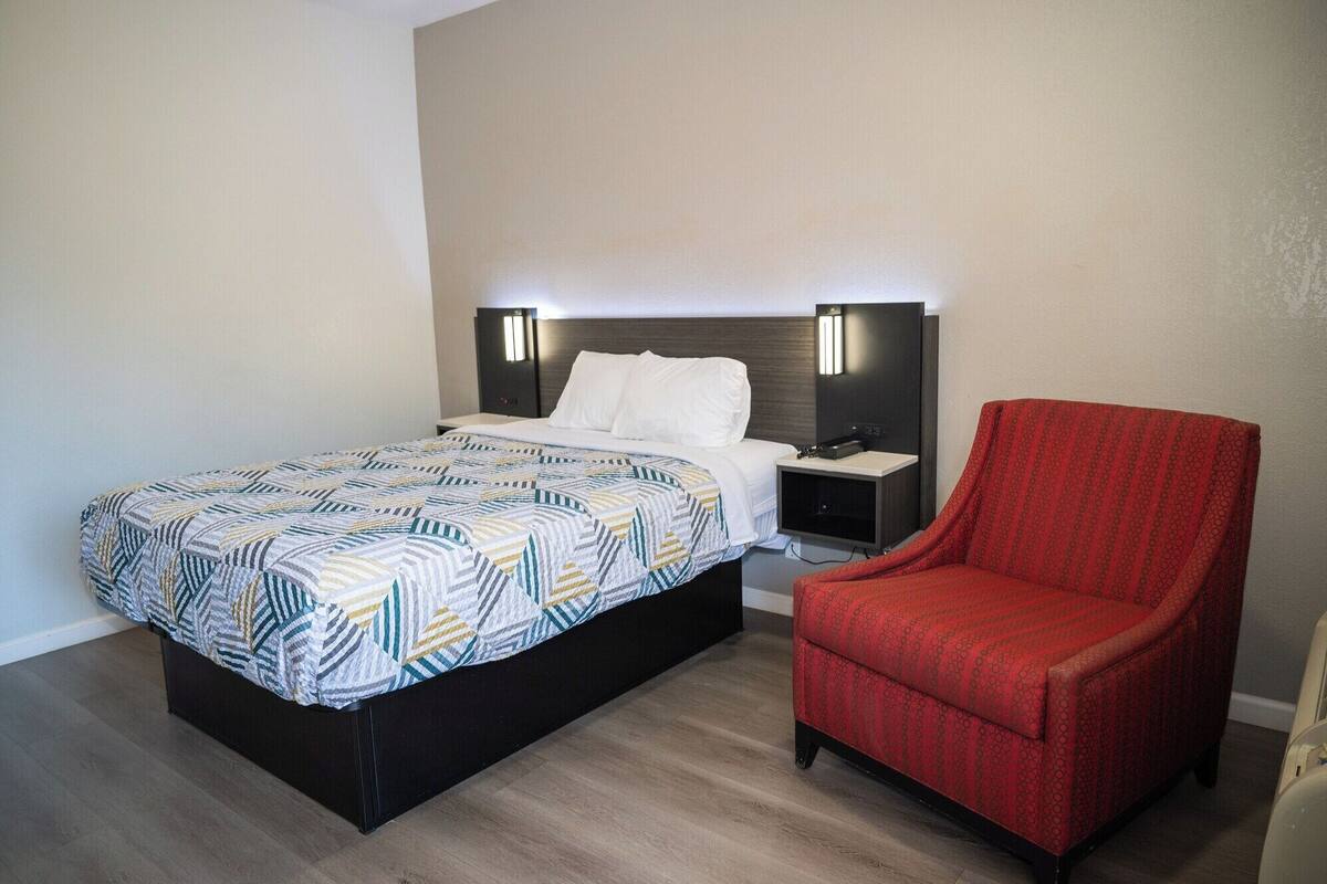 A hotel room with a bed, a nightstand, a red armchair, and a wall-mounted lamp.