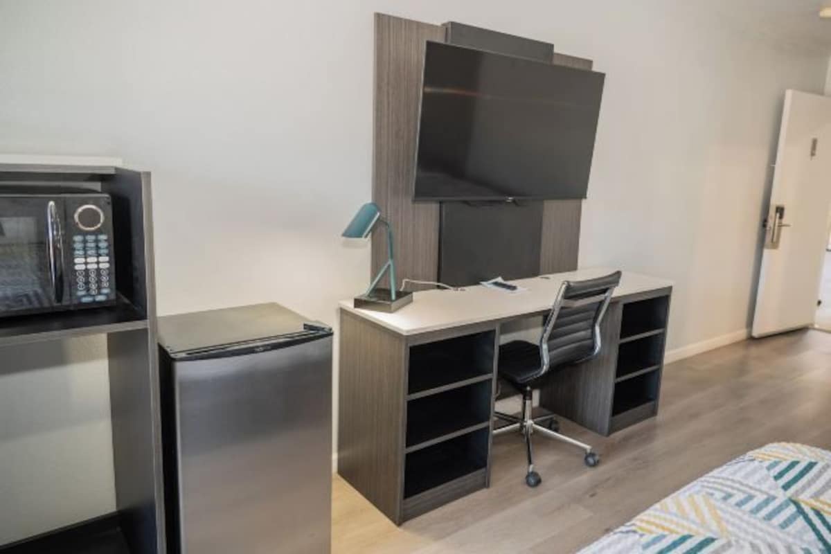 Habitación compacta con kitchenette, un escritorio y silla, un televisor de pantalla plana sobre el escritorio, y una cama con ropa de cama estampada.