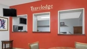 A Travelodge room with a red wall, a round wooden table, two chairs, a TV mounted on the wall, and a framed picture.