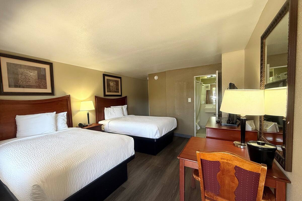 A hotel room with two beds, a desk, a chair, a mirror, and a bathroom visible through an open door.