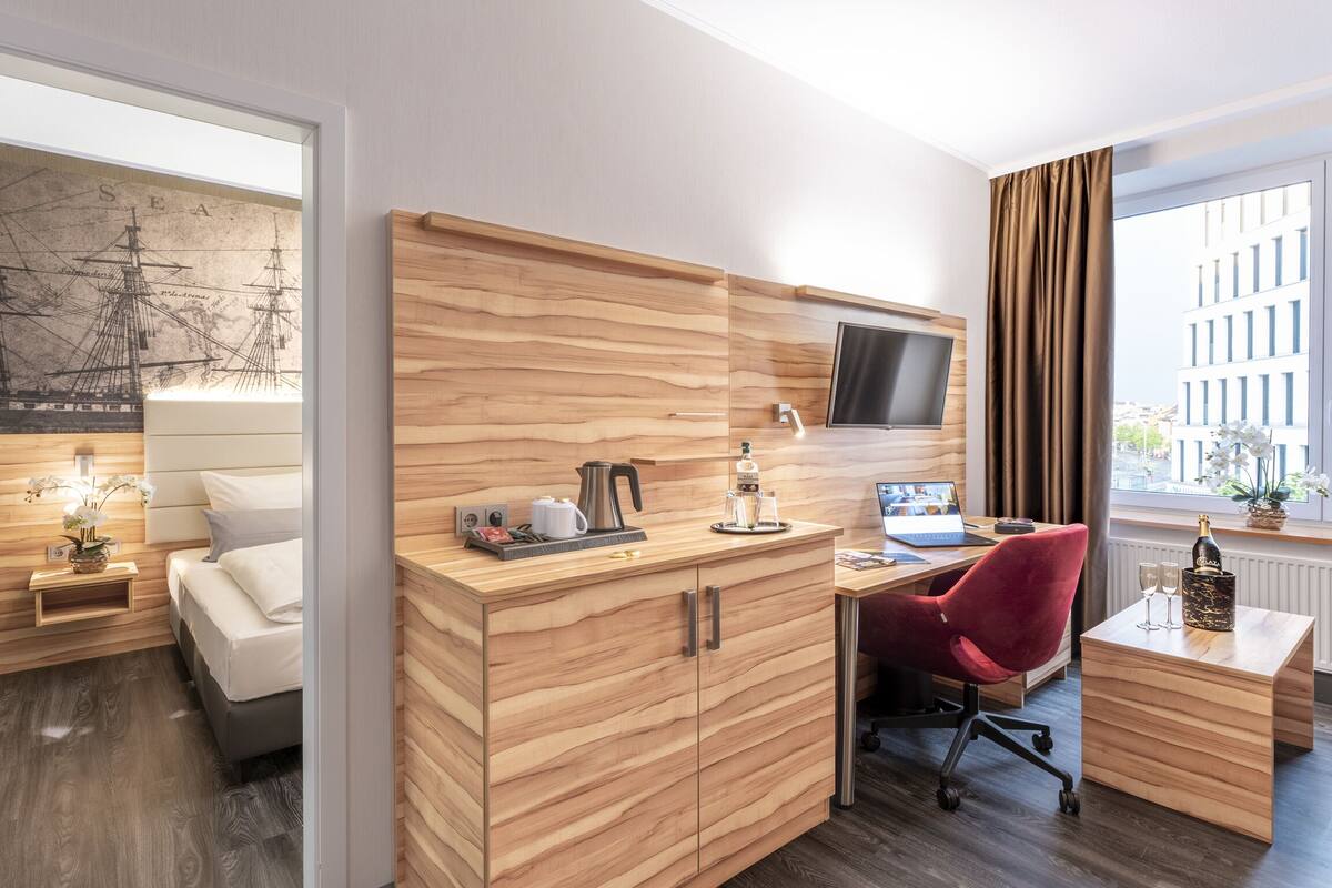 Una camera d'albergo moderna con una scrivania in legno, una sedia rossa, una TV a parete e un'ampia finestra con tende.