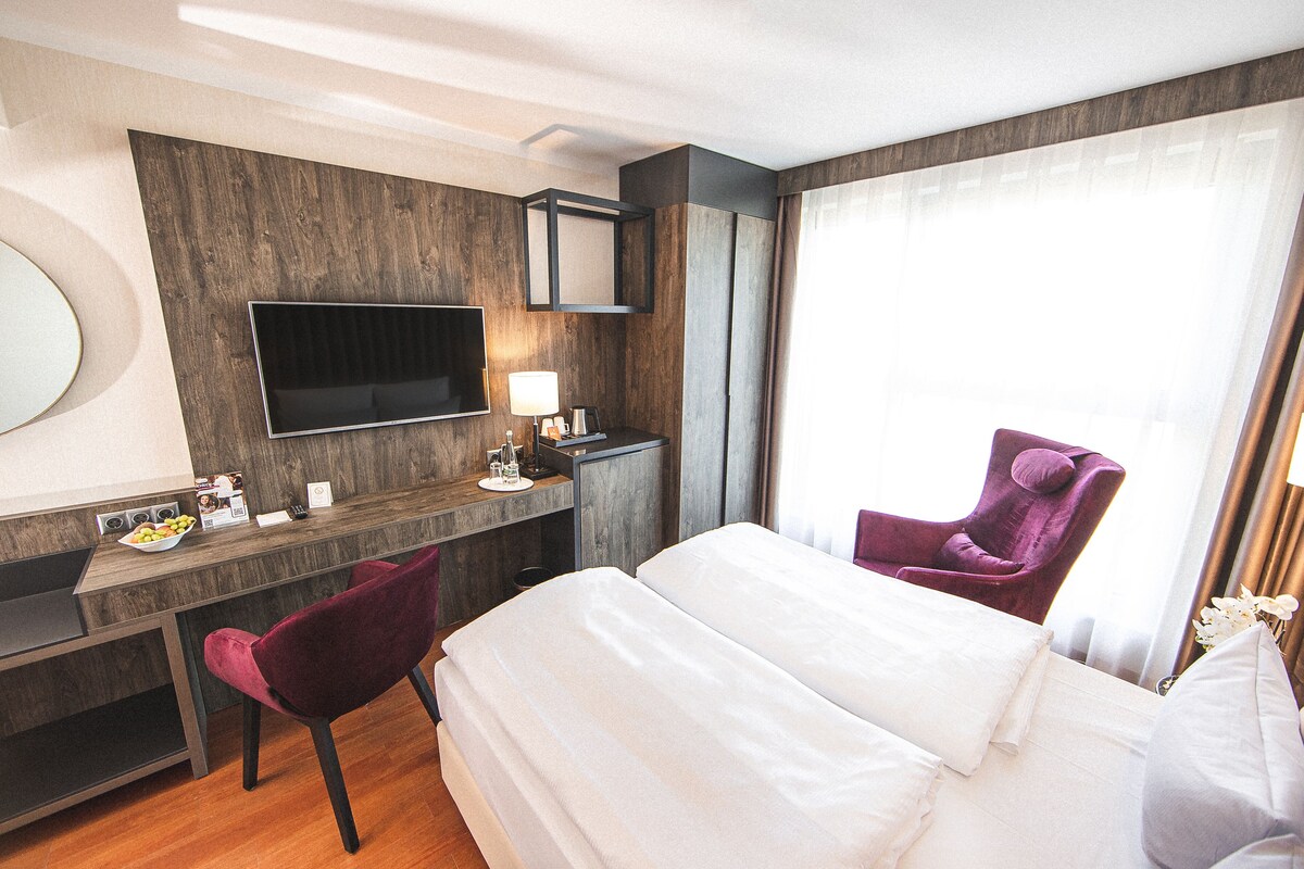Habitación de hotel con cama, escritorio, silla, televisor y ventana con cortinas.