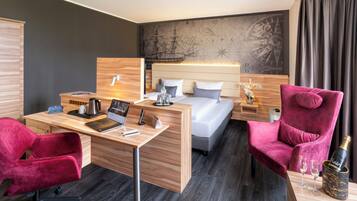 A modern hotel room with a wooden desk, a bed with white linens, a red velvet armchair, and a large map on the wall.