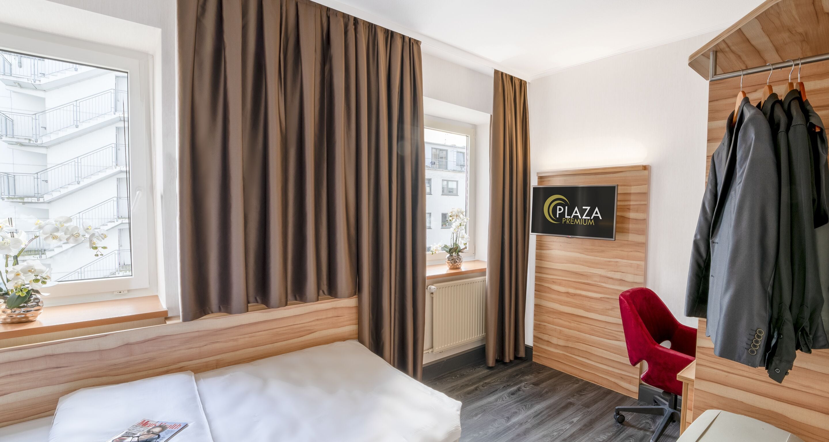A hotel room with a bed, a red chair, a coat rack with clothes, a sign reading "Plaza Premium", and a window with curtains.