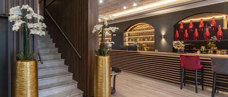 Un bar moderno con mostrador de madera, botellas rojas y una escalera con un jarrón dorado.