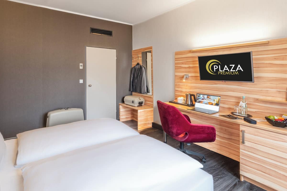 Una camera d'albergo con un letto, una scrivania con un computer, una sedia, uno specchio e un cartello con su scritto 'Plaza Premium'.