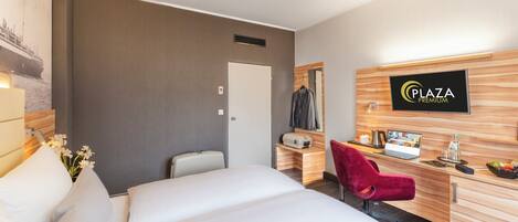 Una camera d'albergo con un letto, una scrivania con un computer, una sedia, uno specchio e un cartello con su scritto 'Plaza Premium'.