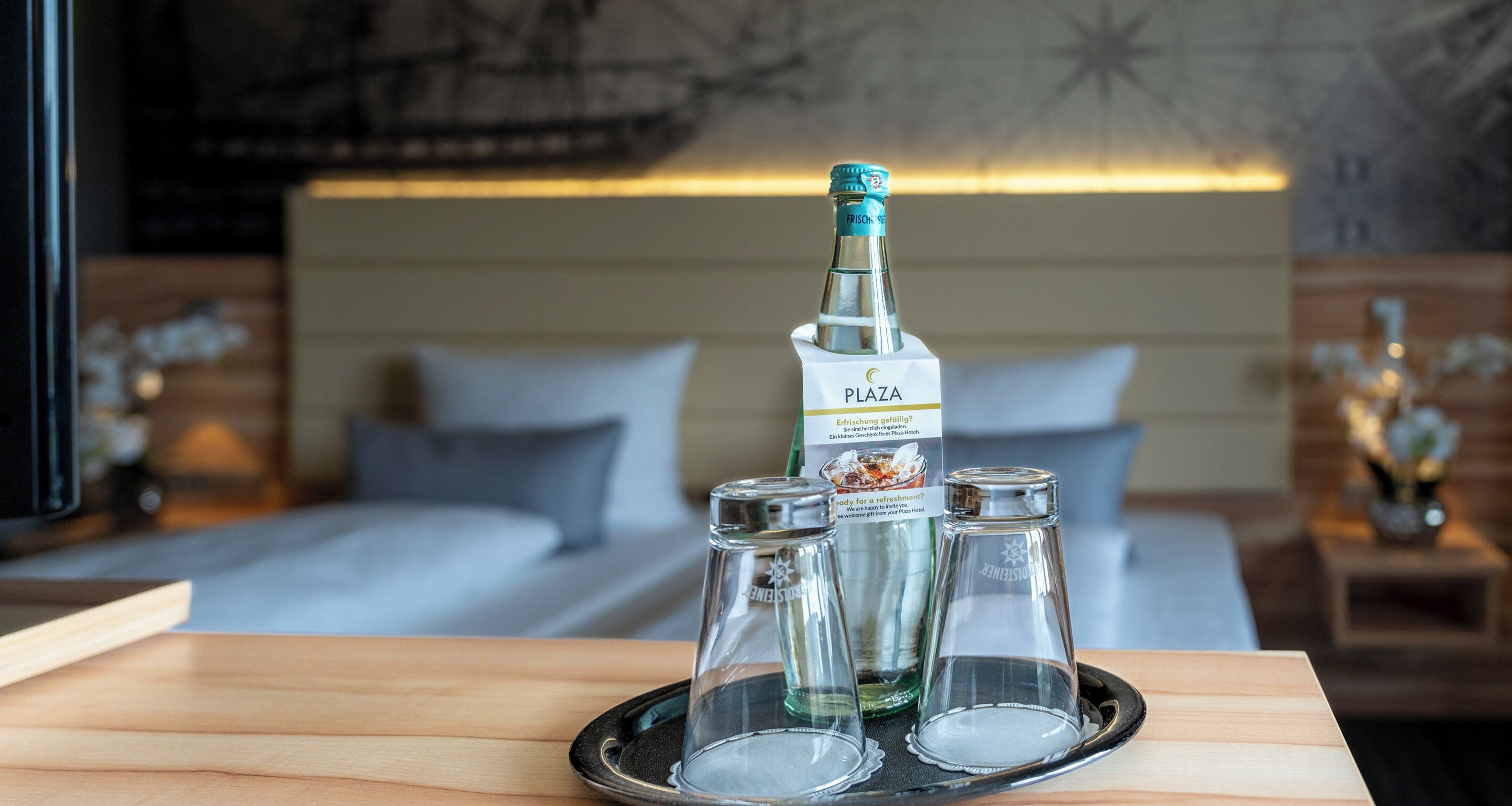 A hotel room with a bed, a bedside table with a tray holding a bottle and two glasses, and a wall with a star pattern.