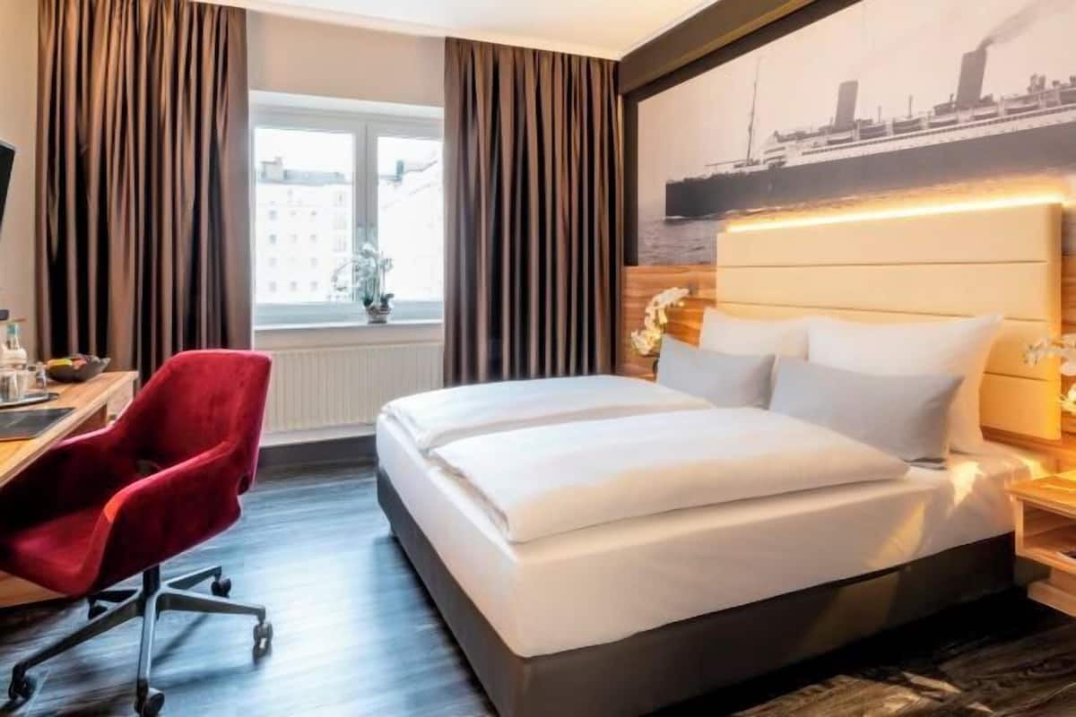 A hotel room with a bed, a desk, a red chair, a window with curtains, and a cityscape painting on the wall.