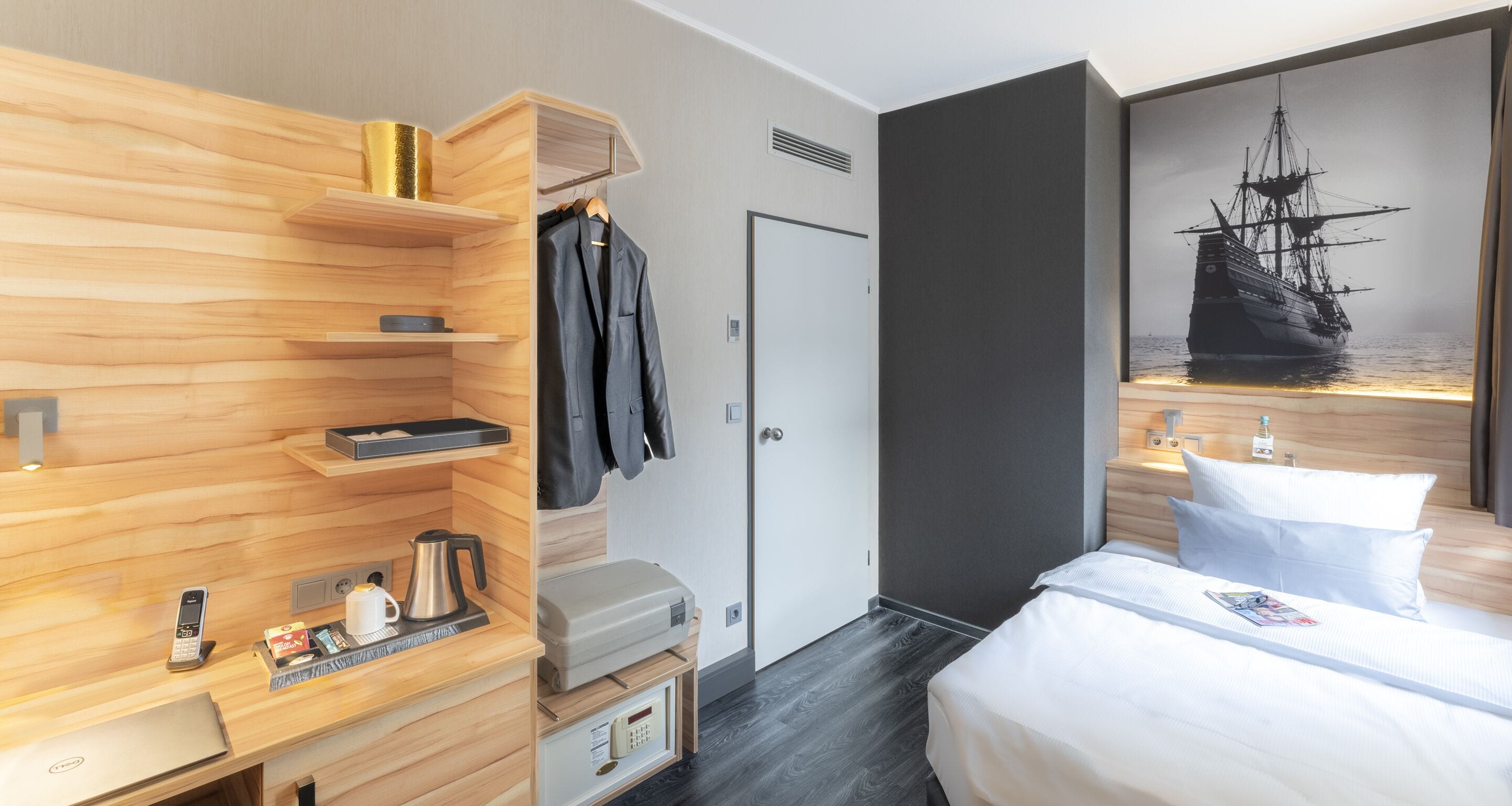 Quarto de hotel com guarda-roupa de madeira, cama com roupa de cama branca, pintura de um navio e pequena mesa com chaleira e telefone.