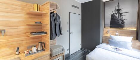 A hotel room with a wooden wardrobe, a bed with white linens, a painting of a ship, and a small table with a kettle and a phone.
