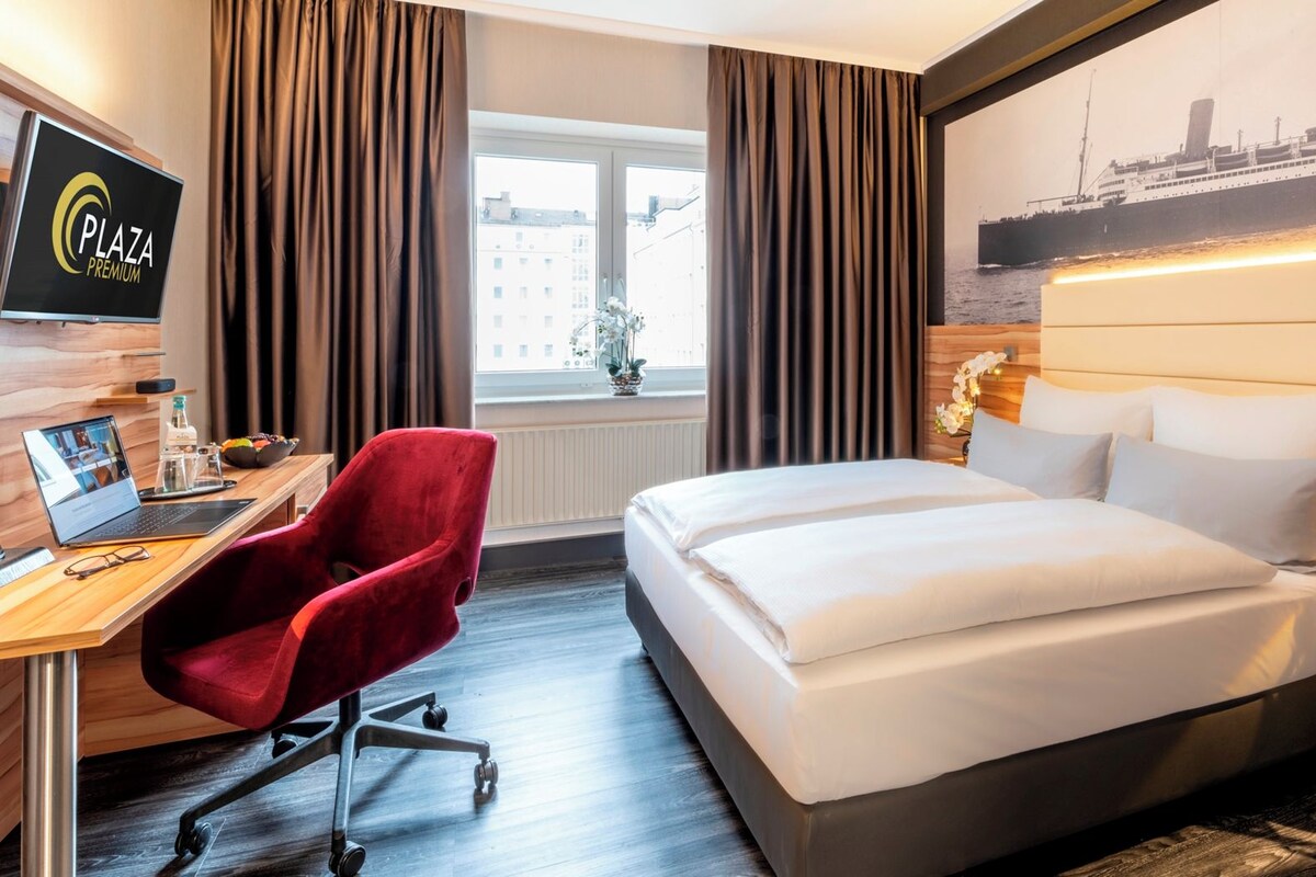 Habitación de hotel con cama, escritorio con una computadora portátil, una silla roja, mesita de noche con lámpara y un ventanal con cortinas.