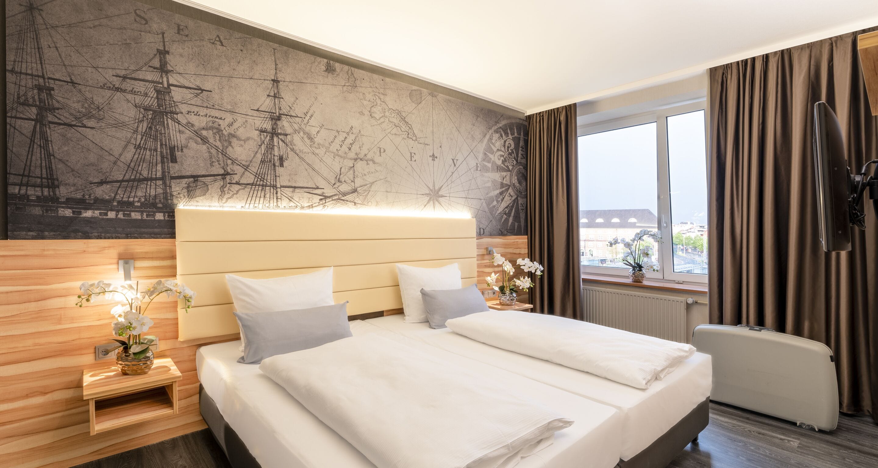 Habitación de hotel con una cama grande, una mesita de noche con un florero y un cabecero de madera, además de un amplio ventanal con cortinas.