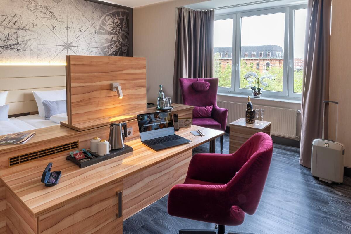 Una camera d'albergo con una scrivania in legno, una sedia viola, un letto con lenzuola bianche, una mappa appesa al muro e una finestra con vista sugli edifici.