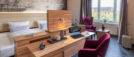 A hotel room with a wooden desk, a purple chair, a bed with white linens, a map on the wall, and a window with a view of buildings.