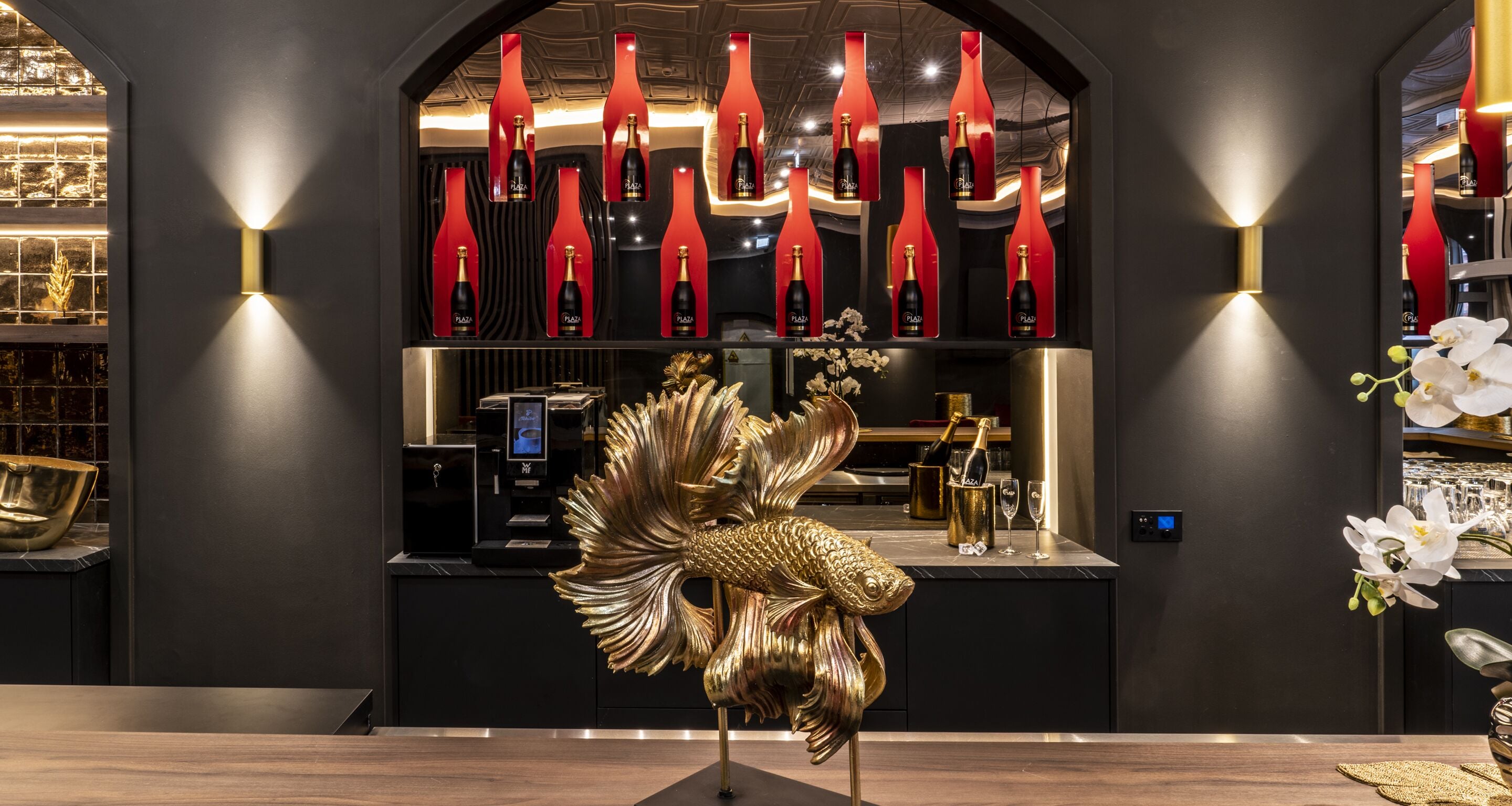 A modern interior with a central display of champagne bottles, a golden fish sculpture, and a bar area.